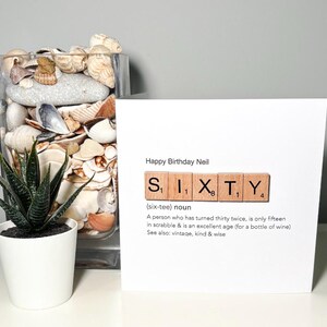 May include: A birthday card with the words "Happy Birthday Neil" and the word "SIXTY" spelled out with Scrabble tiles. The card includes a definition of sixty. A small potted plant and a glass vase filled with shells are also in the image.