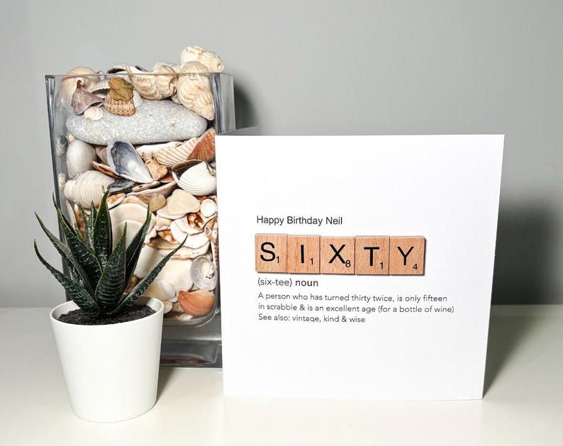 May include: A birthday card with the words "Happy Birthday Neil" and the word "SIXTY" spelled out with Scrabble tiles. The card includes a definition of sixty. A small potted plant and a glass vase filled with shells are also in the image.