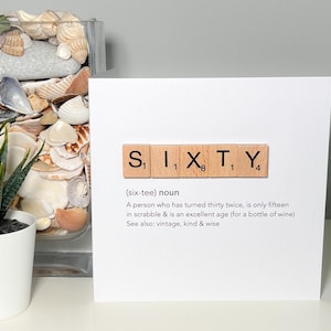May include: A white greeting card with the word "SIXTY" spelled out in wooden Scrabble tiles. The card includes a definition of the word and related terms. A succulent plant in a white pot and a glass jar filled with seashells are in the background.