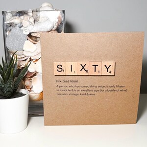 May include: A brown card with the word "SIXTY" spelled out in Scrabble tiles. The card includes a definition of the word "sixty" and is next to a potted succulent and a glass jar filled with seashells.