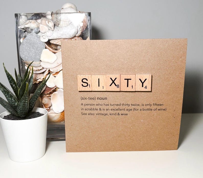 May include: A brown card with the word "SIXTY" spelled out in Scrabble tiles. The card includes a definition of the word "sixty" and is next to a potted succulent and a glass jar filled with seashells.