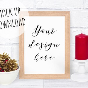 Puede incluir: Un marco blanco con un borde de madera se encuentra sobre una pared de ladrillo blanco. El marco tiene un fondo blanco con el texto "Your design here" escrito en cursiva negra. Una vela roja se encuentra sobre un portavelas blanco a la derecha del marco. Un tazón de piñas doradas y bayas rojas se encuentra a la izquierda del marco.