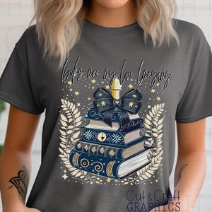Könnte beinhalten: Ein graues T-Shirt mit einem Stapel Bücher, einer Kerze und einer Schleife. Der Text "Books are my love language" ist auf dem T-Shirt aufgedruckt.