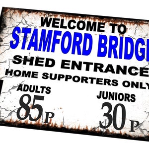 May include: A white sign with black and blue text that reads "Welcome to Stamford Bridge Shed Entrance Home Supporters Only Adults 85p Juniors 30p". The sign has a distressed, rusty look and arrows pointing down on either side.