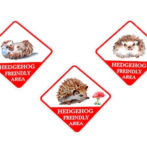 May include: Three red diamond-shaped signs with white borders and the text "Hedgehog Freindly Area" in white. Each sign features a watercolour illustration of a hedgehog.