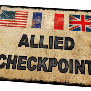 Könnte beinhalten: Ein Schild im Vintage-Stil mit dem Text "Allied Checkpoint" in schwarzen Buchstaben auf beigefarbenem Hintergrund. Das Schild zeigt die Flaggen der Vereinigten Staaten, Frankreichs und Großbritanniens.