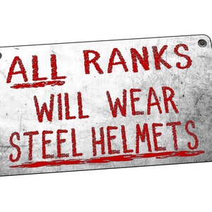Op de afbeelding: Een wit metalen bord met rode tekst die luidt "ALL RANKS WILL WEAR STEEL HELMETS". Het bord heeft een verweerde, versleten uitstraling.