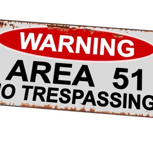 Op de afbeelding: Een roestig metalen bord met een rood ovaal waarschuwingsbord waarop "WARNING" in witte letters staat. Het bord zegt "AREA 51 NO TRESPASSING" in zwarte letters.