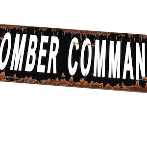Pode incluir: Uma placa de metal retangular com as palavras "BOMBER COMMAND" em letras brancas e em negrito sobre um fundo preto. A placa tem uma aparência desgastada e enferrujada, sugerindo idade ou intemperismo.