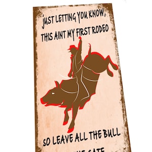 Könnte beinhalten: Ein Schild im Vintage-Stil mit braunem Hintergrund und weißem Text. Das Schild zeigt eine Silhouette eines Cowboys, der einen sich aufbäumenden Stier reitet. Der Text lautet "Just letting you know, this ain't my first rodeo. So leave all the bull at the gate."