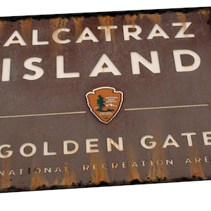 May include: A rusty brown metal sign with white text that reads "Alcatraz Island Golden Gate National Recreation Area". The sign also features a brown and green National Park Service emblem.