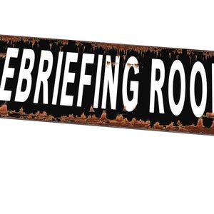 Op de afbeelding: Een roestig zwart metalen bord met witte letters met de tekst "DEBRIEFING ROOM".