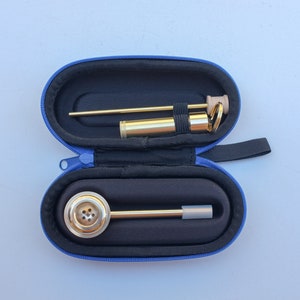 K&ouml;nnte beinhalten: Eine goldfarbene Metall-Rauchr&ouml;hre mit einer runden Schale und einem langen, d&uuml;nnen Schaft. Die Pfeife befindet sich in einem blauen Transportkoffer mit Rei&szlig;verschluss.
