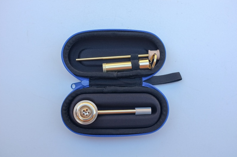 K&ouml;nnte beinhalten: Eine goldfarbene Metall-Rauchr&ouml;hre mit einer runden Schale und einem langen, d&uuml;nnen Schaft. Die Pfeife befindet sich in einem blauen Transportkoffer mit Rei&szlig;verschluss.