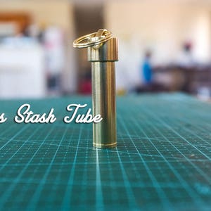 Puede incluir: Un tubo de almacenamiento de latón con un llavero. El tubo está sobre una estera de corte verde.