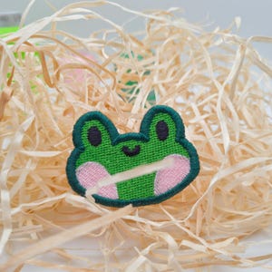 May include: Embroidered green frog patch with pink cheeks and a dark green outline. The frog's eyes are black, and it has a small, curved mouth. The patch is set against a background of light brown straw.