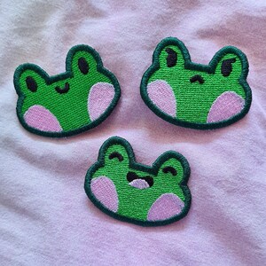 May include: Three green felt frog patches with pink cheeks. The frogs have different facial expressions.