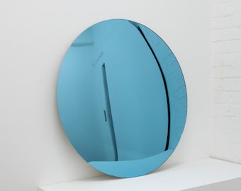 Puede incluir: Un espejo redondo de color azul con un reflejo ligeramente distorsionado de una pared blanca y una puerta. El espejo está sobre una superficie blanca.