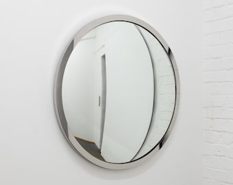 Pode incluir: Um espelho redondo com moldura prateada e uma reflexão ligeiramente distorcida. O espelho está pendurado em uma parede branca.