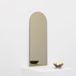 May include: A tall, arched mirror with a gold-toned frame and a reflective surface. The mirror is standing on a white surface with a small, gold-toned bowl in front of it.
