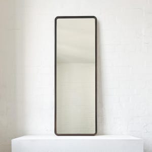 May include: A tall, rectangular mirror with a dark brown frame. The mirror is placed on a white surface against a white brick wall. The frame has rounded corners, and the mirror's surface reflects the surrounding environment.