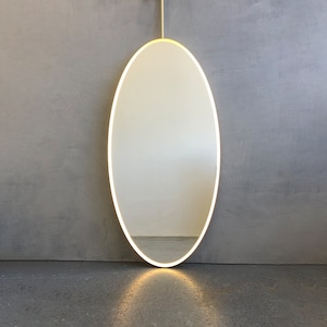 May include: A large oval mirror with a thin, white, illuminated border. The mirror is hanging on a gray wall.