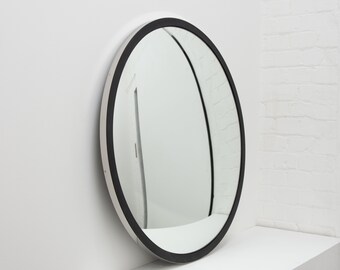 Pode incluir: Um espelho redondo com moldura preta e borda externa branca. O espelho reflete uma porta branca e uma parede de tijolos. O design é minimalista e moderno.