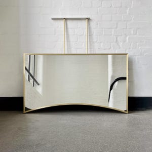 Pode incluir: Um espelho grande e curvo com uma moldura dourada. O espelho está pendurado em uma parede de tijolo branco.