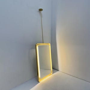 Pode incluir: Um espelho com moldura dourada e uma luz branca quente em torno do perímetro. O espelho está pendurado no teto com uma haste dourada.