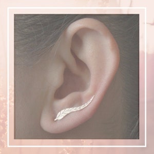 Może przedstawiać: Srebrny kolczyk w kształcie liścia. Kolczyk jest noszony w górnej części ucha, a wzór liścia rozciąga się na zewnątrz i od ucha.