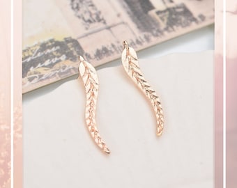 May include: Two rose gold leaf-shaped earrings. Each earring has a curved, elongated design with detailed veining. The earrings are set against a soft, neutral background, suggesting a focus on jewelry and accessories.