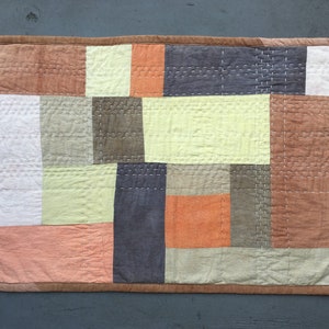 Puede incluir: Una colcha de retazos con cuadrados de varios colores y texturas, incluyendo marrón, naranja, beige y blanco. La colcha está cosida con hilo blanco y tiene un borde marrón.