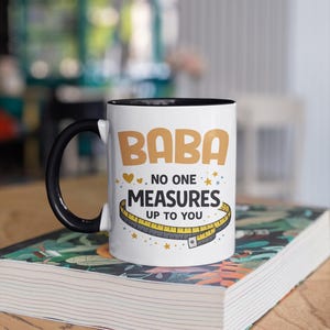 May include: A white ceramic mug with a black handle and interior. The mug features the text "BABA" in large tan letters, followed by "NO ONE MEASURES UP TO YOU" in black, accompanied by a yellow measuring tape graphic and small stars.