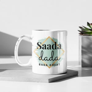 May include: A white ceramic mug featuring a central design with a light green brushstroke background, a gold diamond outline, and black text that reads Saada dada BADA GREAT. The mug has a curved handle on the left side and a smooth, glossy finish.