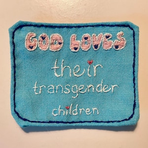 Op de afbeelding: Een rechthoekige, turquoise stoffen patch met geborduurde tekst. De zin "GOD LOVES" is gestikt in roze draad met blauwe hartaccenten. Daaronder staat in witte draad "their transgender children", met rode hartaccenten.