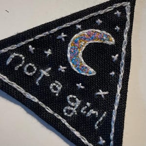 May include: Black triangular patch with white embroidered text and accents. The text reads "Not a Girl" in cursive. A crescent moon with colorful embroidery is in the upper right corner. The patch is bordered with white stitching.