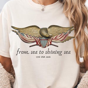 May include: Cream-colored t-shirt featuring a detailed illustration of an eagle with outstretched wings, and a banner with red and white stripes. The text "from sea to shining sea" and "1776 USA 2026" are printed below the eagle. The shirt has a classic crew neck.