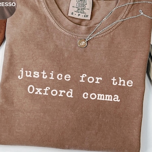 Könnte beinhalten: Ein espressofarbenes T-Shirt mit Rundhalsausschnitt und kurzen Ärmeln. Das Shirt zeigt den weißen Aufdruck 'justice for the Oxford comma' auf der Brust. Eine silberfarbene Kette mit einem kleinen runden Anhänger liegt auf dem Stoff.