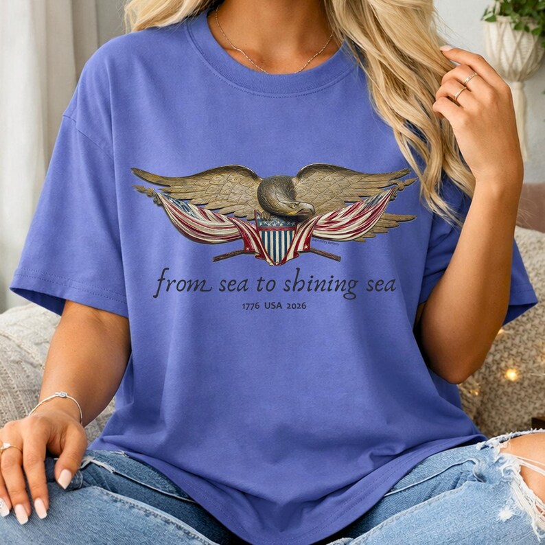 May include: A periwinkle blue t-shirt featuring a detailed illustration of an eagle with outstretched wings, holding a shield with red, white, and blue stripes. The text "from sea to shining sea" and "1776 USA 2026" are printed below the eagle.