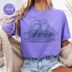 Pu&ograve; includere: T-shirt viola con un'illustrazione vintage di una barca a vela e la scritta "Swim" in corsivo. La maglietta ha maniche corte e una vestibilit&agrave; rilassata, perfetta per l'abbigliamento casual.