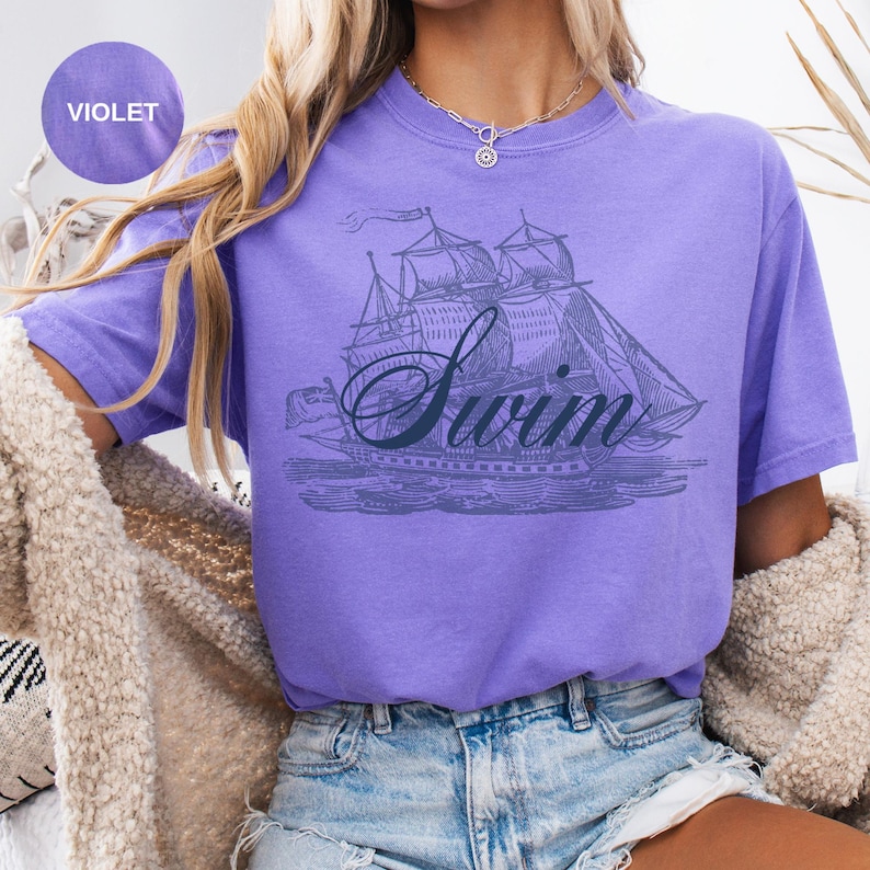 Pu&ograve; includere: T-shirt viola con un'illustrazione vintage di una barca a vela e la scritta "Swim" in corsivo. La maglietta ha maniche corte e una vestibilit&agrave; rilassata, perfetta per l'abbigliamento casual.