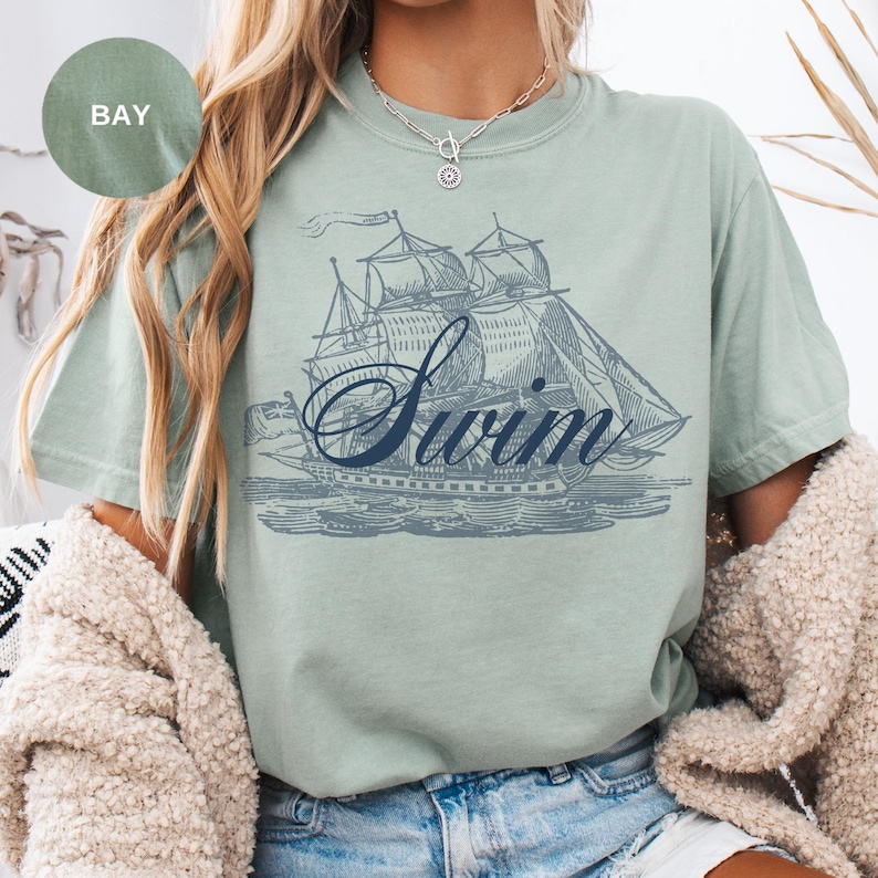 Pu&ograve; includere: T-shirt verde chiaro con un'illustrazione blu di una barca a vela e la parola "Swim" in corsivo. La maglietta ha maniche corte e una vestibilit&agrave; rilassata. La parola "BAY" &egrave; in un cerchio in alto a sinistra.