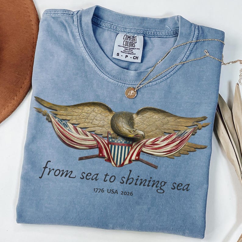 May include: A light blue Comfort Colors t-shirt featuring an eagle design with American flag elements. The shirt has the text "from sea to shining sea" and "1776 USA 2026". A gold necklace with a pendant rests on the shirt.