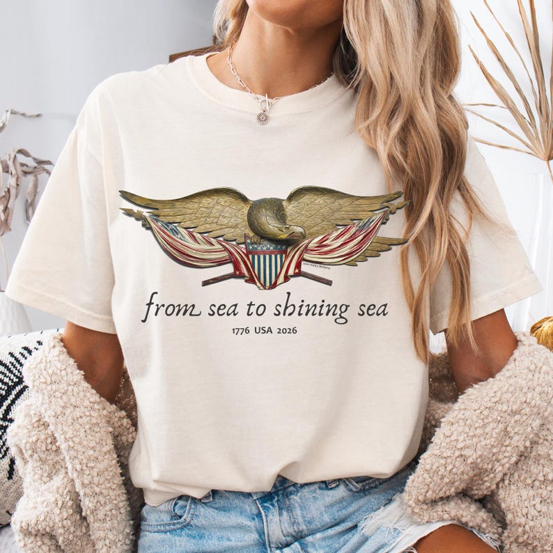 May include: Cream-colored t-shirt featuring a detailed illustration of an eagle with outstretched wings, and a banner with red and white stripes. The text "from sea to shining sea" and "1776 USA 2026" are printed below the eagle. The shirt has a classic crew neck.