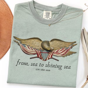 May include: A sage green t-shirt with a detailed eagle graphic, American flag accents, and the phrase "from sea to shining sea." The shirt includes the text "1776 USA 2026" and a small tag that says "Comfort Colors". A gold necklace with a star pendant is also visible.