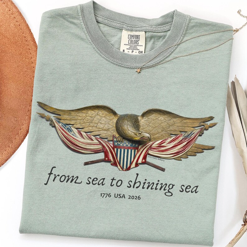 May include: A sage green t-shirt with a detailed eagle graphic, American flag accents, and the phrase "from sea to shining sea." The shirt includes the text "1776 USA 2026" and a small tag that says "Comfort Colors". A gold necklace with a star pendant is also visible.