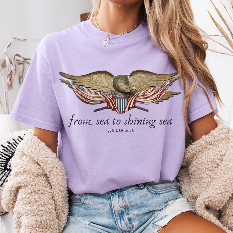 May include: A light purple t-shirt featuring a detailed illustration of an eagle with outstretched wings, incorporating elements of the American flag. The text "from sea to shining sea" and "1776 USA 2026" are printed below the eagle.