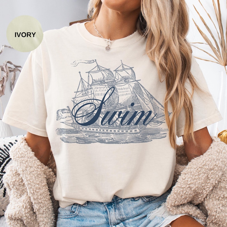 May include: Ivory-colored t-shirt featuring a blue illustration of a sailboat with the word "Swim" in a script font. The shirt has a relaxed fit and short sleeves. The design is reminiscent of vintage nautical themes.