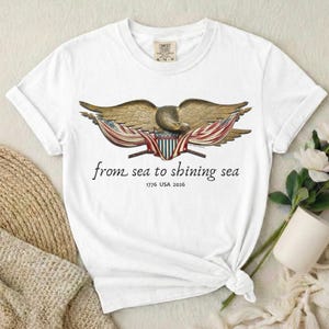 May include: White t-shirt featuring a detailed illustration of a golden eagle with outstretched wings, holding American flags. The text "from sea to shining sea" and "1776 USA 2026" are printed below the eagle. The shirt is made of a soft material.