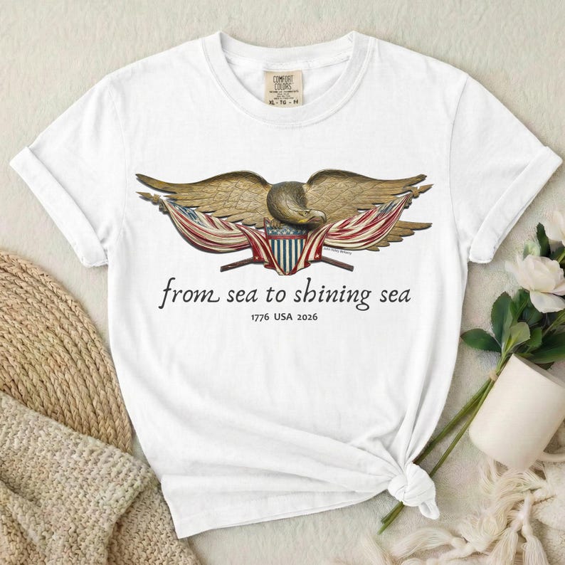 May include: White t-shirt featuring a detailed illustration of a golden eagle with outstretched wings, holding American flags. The text "from sea to shining sea" and "1776 USA 2026" are printed below the eagle. The shirt is made of a soft material.
